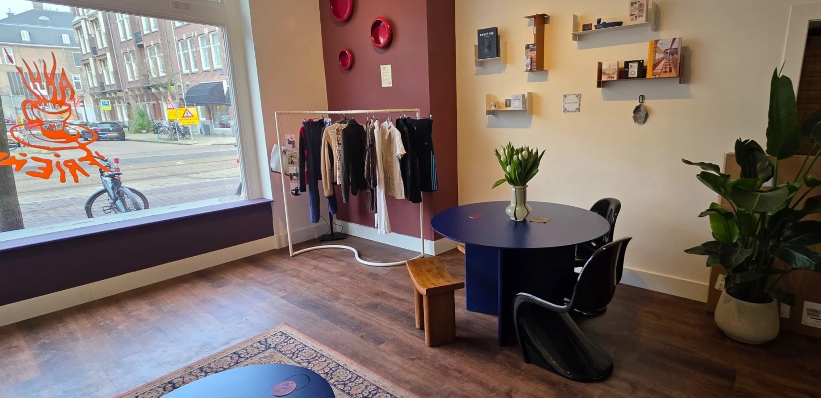 Inside Akhi 'n Friends: the living-room seating area with a rug, shelves of books and a rack of clothing beside the coffee bar